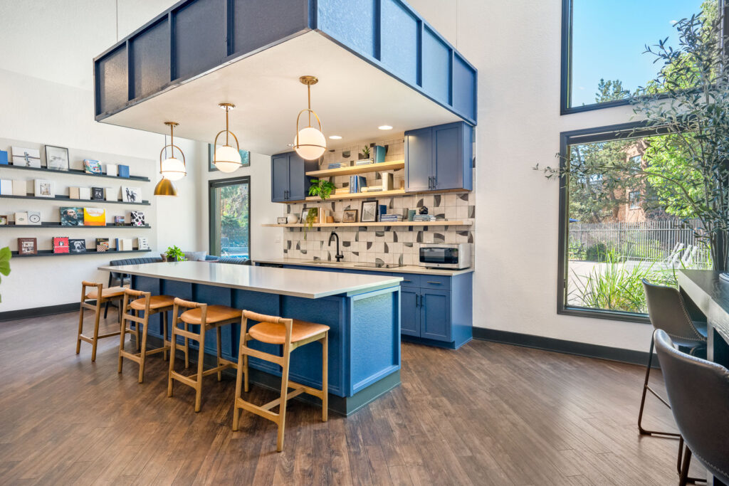 Kitchen and lounge area with blue cabinets, large center island with five wooden stools. Open shelves hold decor, books and plants. Large windows for natural light and view of pool deck.