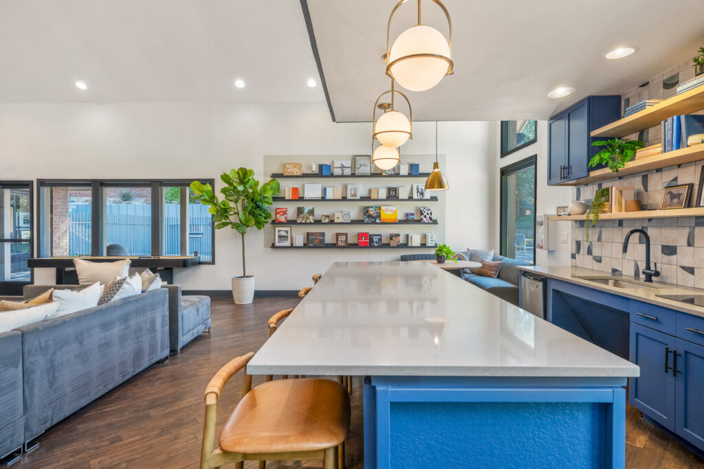 Large center island with wooden stools and a wall with mounted shelving with books and album covers. Shuffleboard table adjacent to couch seating across from the kitchen space.