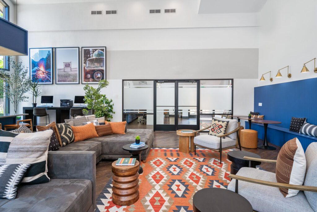 Large communal sitting space with chairs and sectional couch facing a wall mounted television above an electric fireplace. Back wall has double door access to a front office space, adjacent to that is a computer desk with printer.