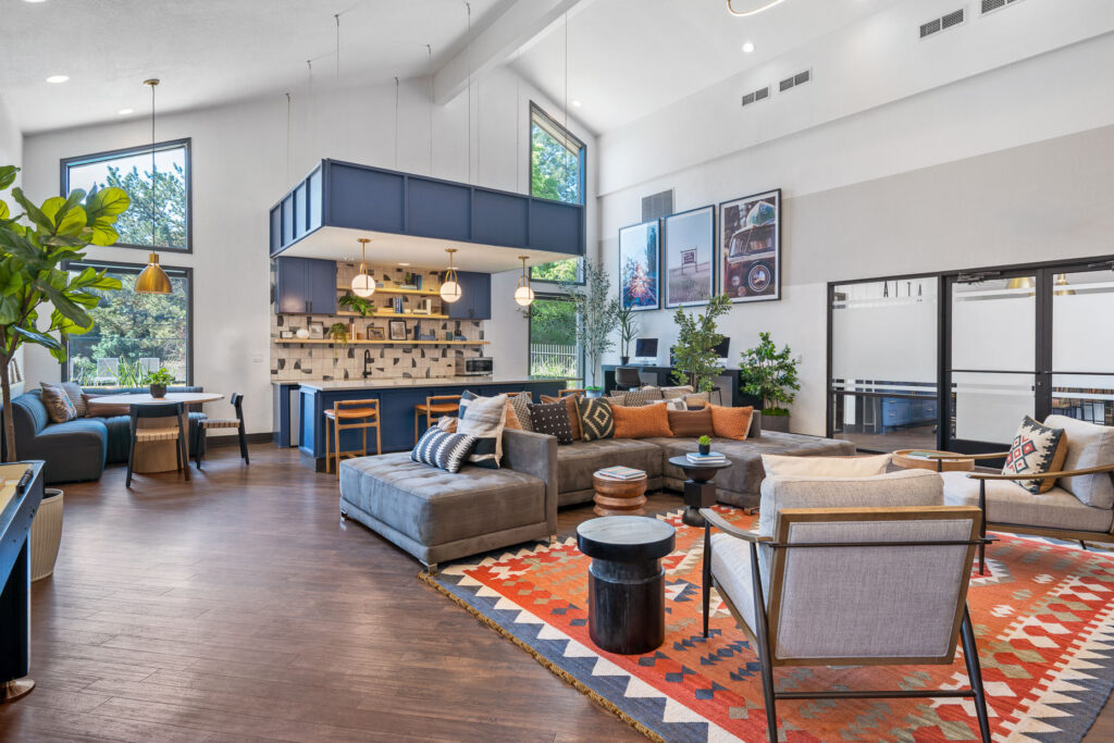 Community lounge and kitchen area with large sectional sofa and armchairs. Kitchen space has blue cabinets, large island with bar stools and open shelving. There are computers and framed artwork along the far wall.