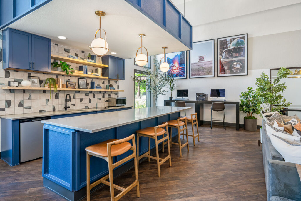 Communal kitchen space with microwave, dishwasher, mini fridge and sink. Kitchen cabinets are blue with a large center island and bar stools. The adjacent wall has computers and a printer.