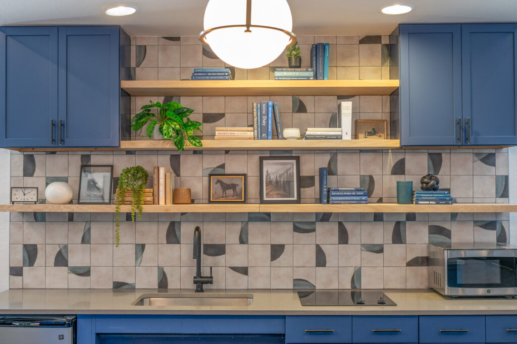 Modern open shelving unit above a communal kitchen space with blue cabinets and wooden shelving. Shelves are decorated with books and art.